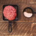 Raw Wagyu ancestral ground beef in a cast iron skillet with a small bowl of spices on a wooden surface.